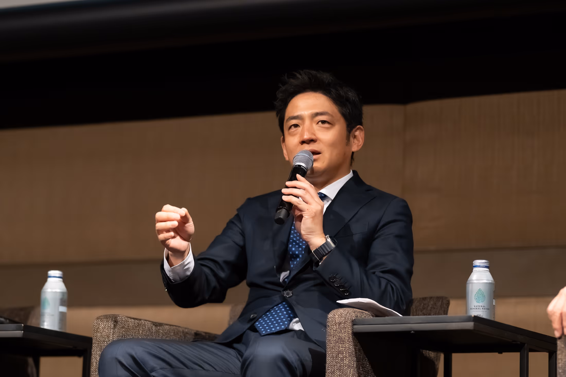 スーツ姿の男性がマイクを持ち、椅子に座って話している。