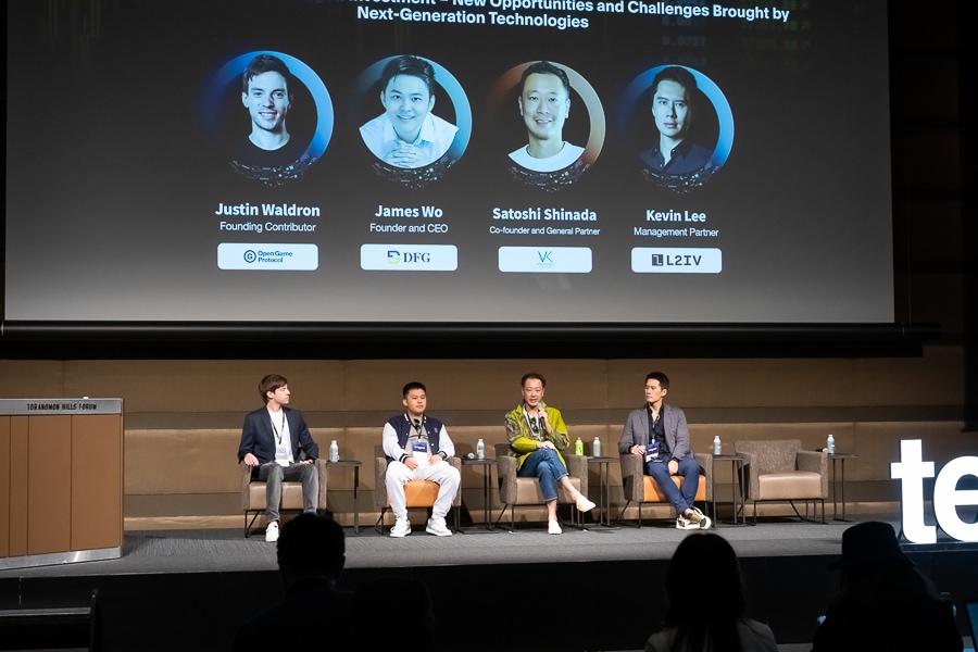 パネル討論中に4人の男性がステージ上の椅子に座っている。背景には、ジャスティン・ウォルドロン、ジェームズ・ウォー、シンナダ・サトシ、ケビン・リーの写真と肩書きがスクリーンに表示されている。