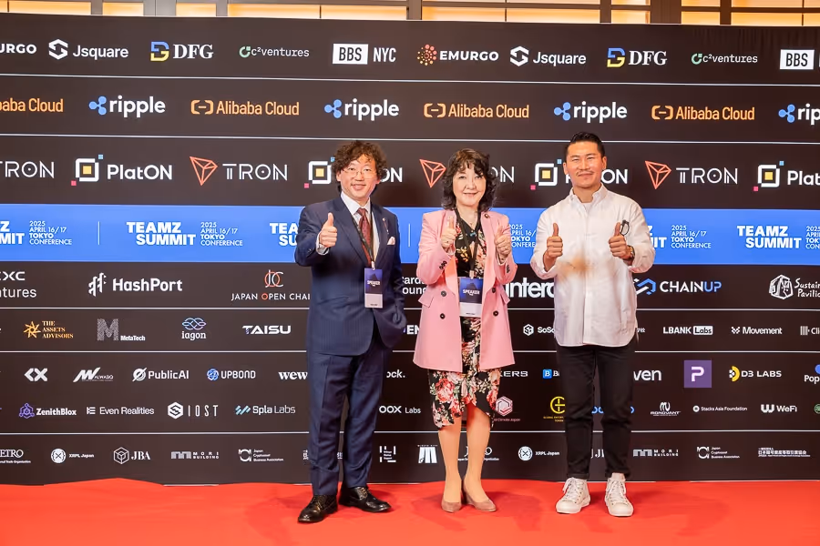 スーツを着た男性、ピンクのジャケットを着た女性、白いシャツの男性が赤いカーペットの上で親指を立てて笑顔でポーズしている。