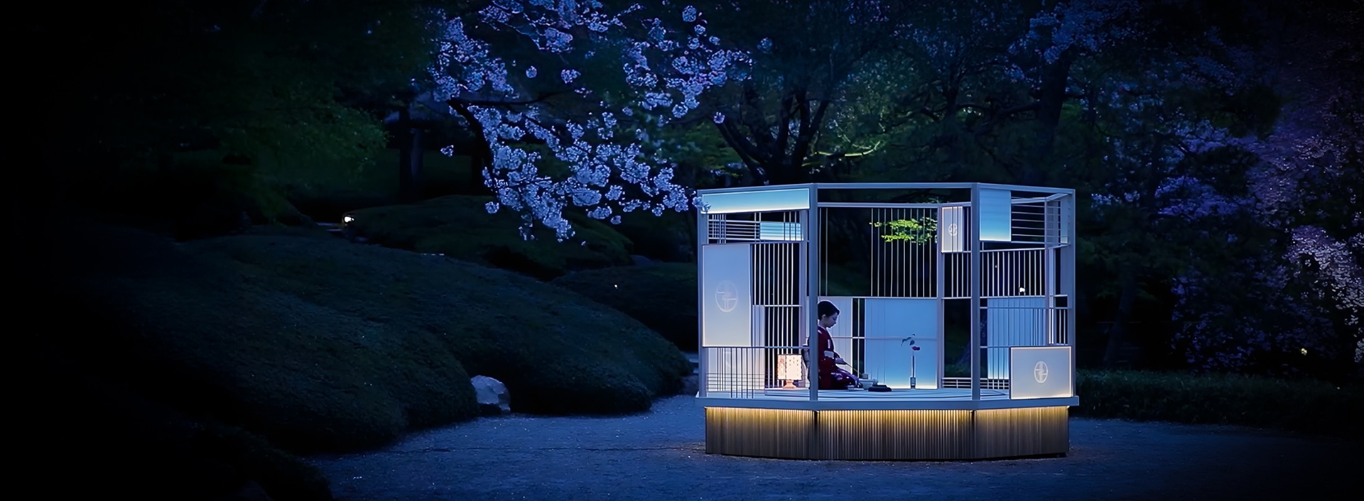 夜の庭園で、桜の木の下に設置された照明付きの和風の囲いの中で座る着物姿の女性。