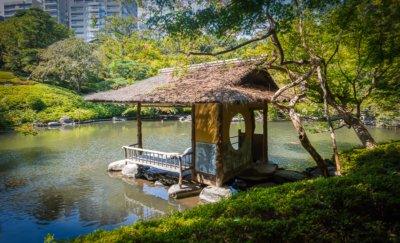 池の上に建てられた藁ぶき屋根の日本風あずまやと周囲の緑豊かな庭園。
