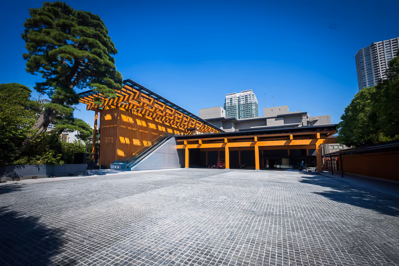 青空の下、現代的な木造の構造が特徴の建物の入口、周囲に高層ビルと緑の木々がある。